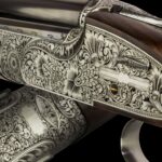 A close-up view of an intricately engraved shotgun, showcasing detailed floral and scrollwork patterns on the metal parts