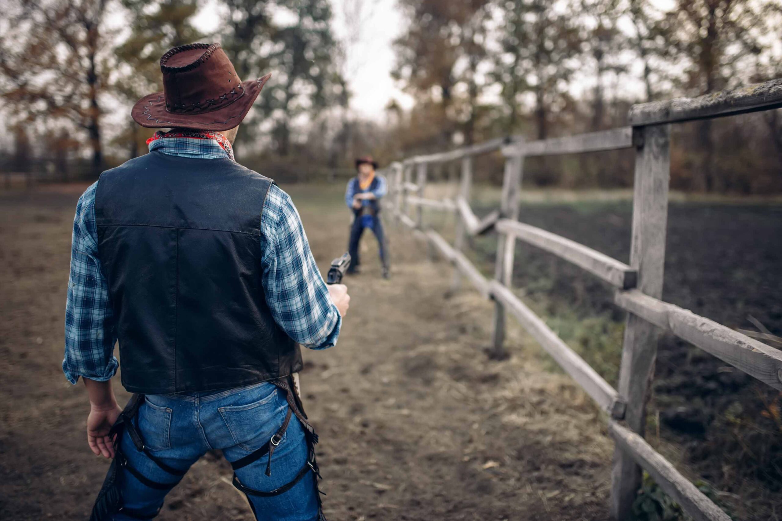 Two cowboys face off in a duel on a dirt path lined with a wooden fence, surrounded by trees.