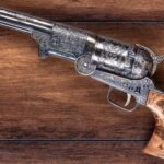 An intricately engraved antique revolver with a wooden grip, displayed on a wooden surface.