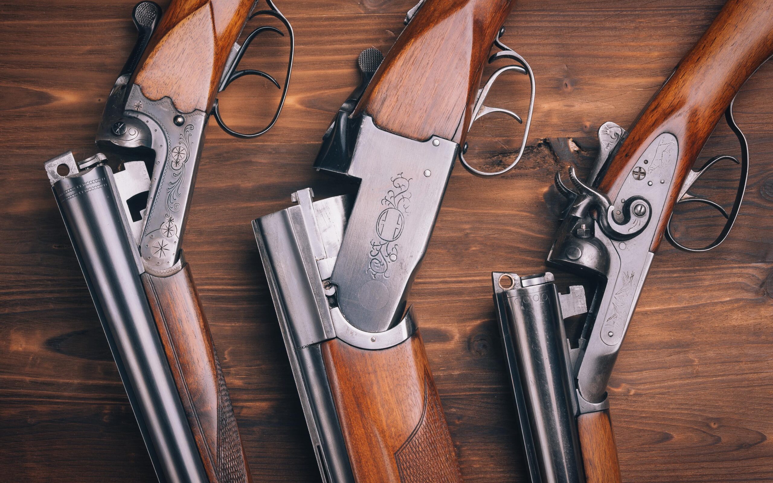 Three vintage double-barreled shotguns with intricate engravings on their metal parts, displayed on a wooden surface.