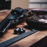 -up of an old-fashioned revolver, ornate powder flask, lead balls, and metal tool on a wooden surface