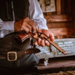 Person in vintage Western-style clothing inspecting a classic revolver indoors, with a green metal chest and poster in the background.
