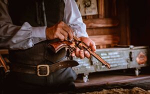 Person in vintage Western-style clothing inspecting a classic revolver indoors, with a green metal chest and poster in the background.
