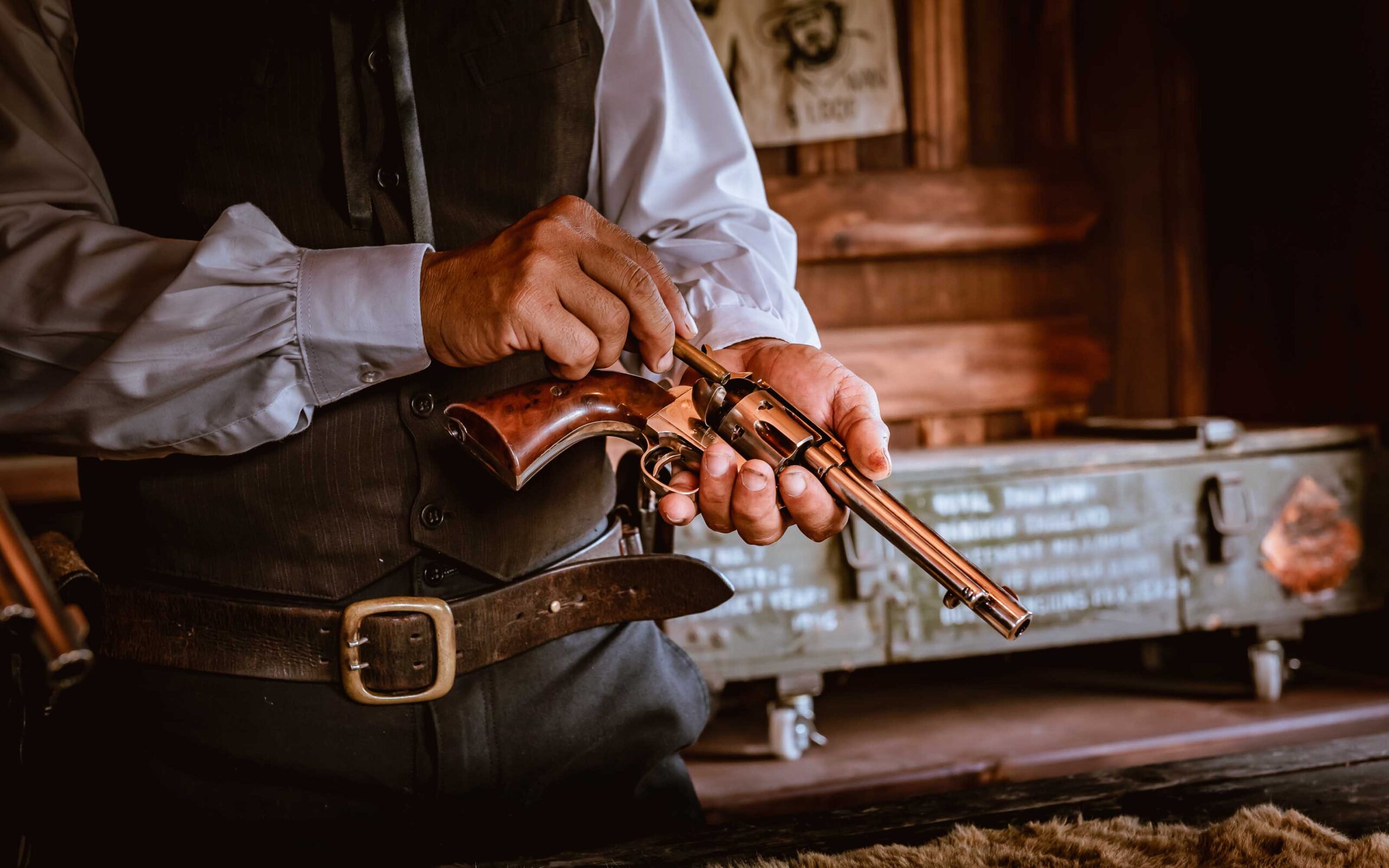 Person in vintage Western-style clothing inspecting a classic revolver indoors, with a green metal chest and poster in the background.