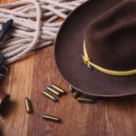 Western-themed items on a wooden surface, including a cowboy hat, revolver, bullets, coiled rope, leather whip, and part of a lever-action rifle.