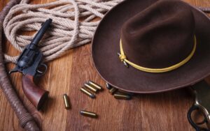 Western-themed items on a wooden surface, including a cowboy hat, revolver, bullets, coiled rope, leather whip, and part of a lever-action rifle.
