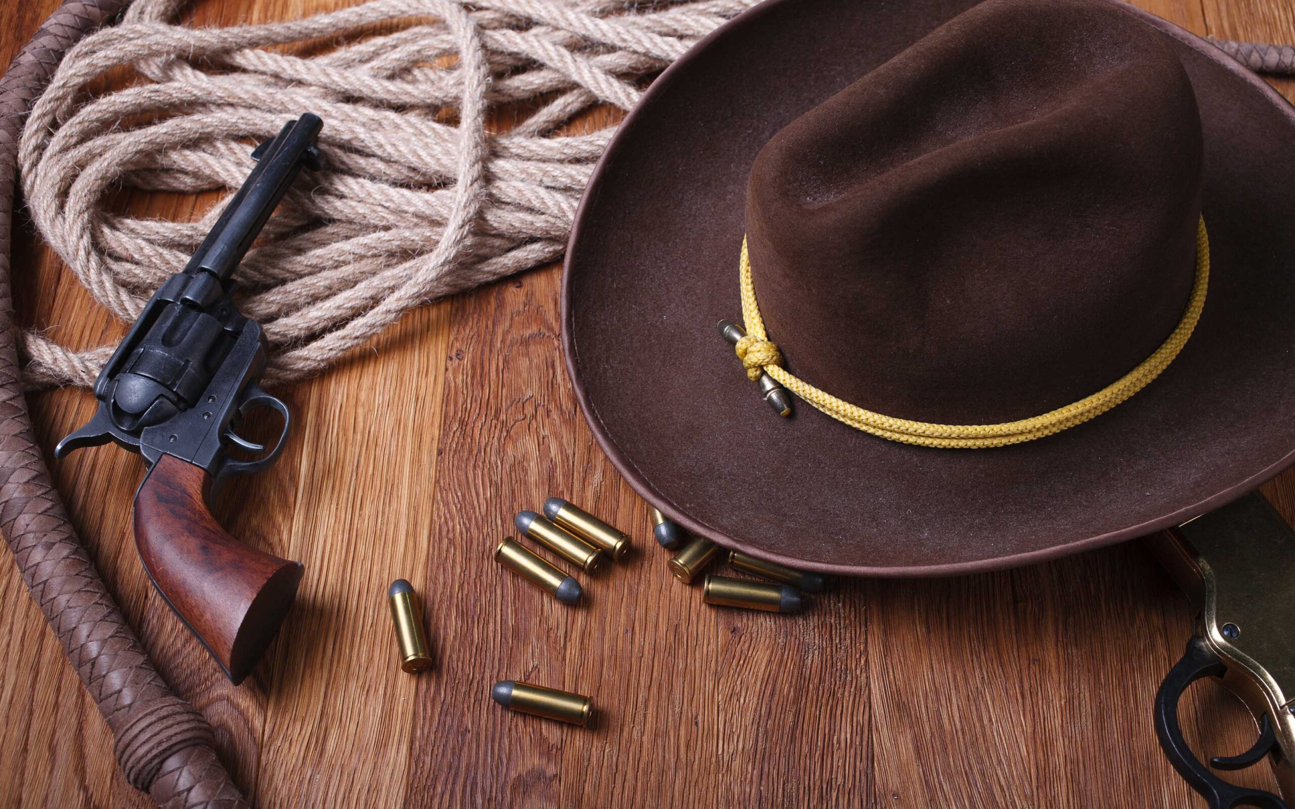 Western-themed items on a wooden surface, including a cowboy hat, revolver, bullets, coiled rope, leather whip, and part of a lever-action rifle.