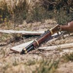 Lever-action rifle with a wooden stock and brass receiver lying on dry grass and broken wooden planks in a rustic outdoor setting