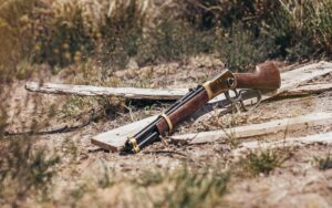 Lever-action rifle with a wooden stock and brass receiver lying on dry grass and broken wooden planks in a rustic outdoor setting