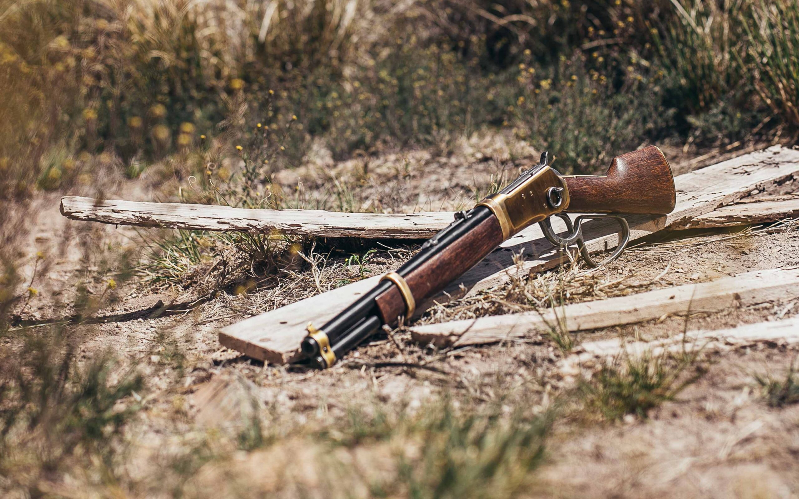 Lever-action rifle with a wooden stock and brass receiver lying on dry grass and broken wooden planks in a rustic outdoor setting