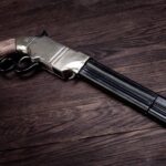 A museum-quality photograph of a vintage replica sawed-off lever-action firearm resting diagonally on a dark, textured wood surface.