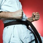 A martial artist with a black belt, wearing a white gi, holds a disciplined fighting stance with clenched fists against a red background.