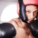 A close-up of a focused young boxer looking at the camera, wearing black boxing gloves and red-and-black sparring headgear featured in a buyer's guide.