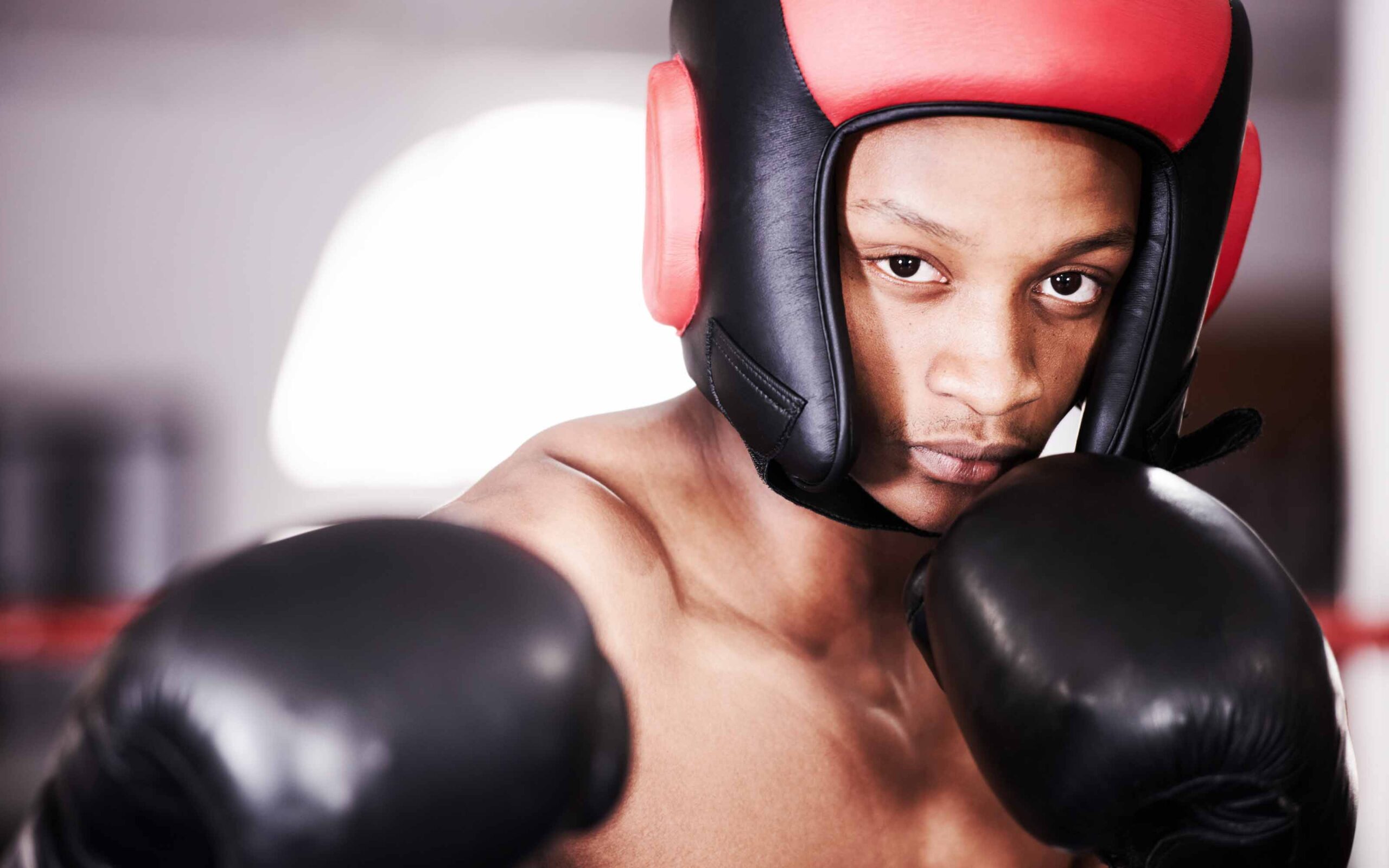 A close-up of a focused young boxer looking at the camera, wearing black boxing gloves and red-and-black sparring headgear featured in a buyer's guide.